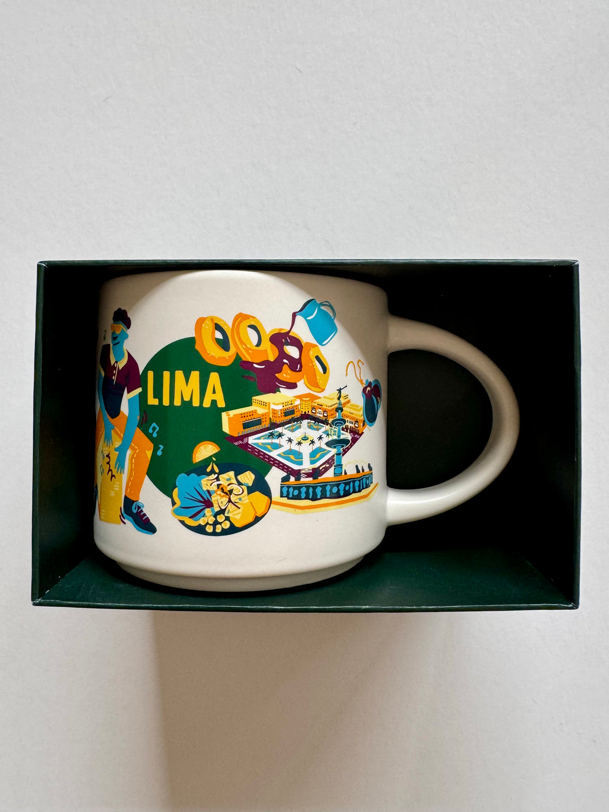 A white ceramic mug with a colorful 'LIMA' travel-themed illustration, presented inside a dark green box on a light gray background.